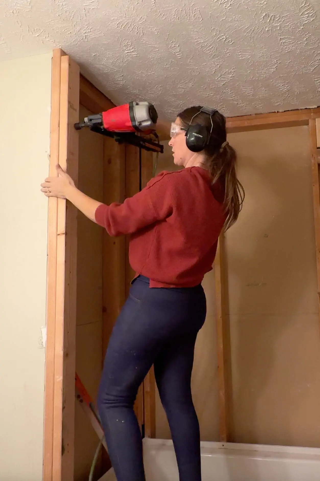 A woman in a red sweater, ear protection, and safety goggles uses a red nail gun to secure framing in place. The bare studs and ceiling are visible as part of the bathroom remodeling process.